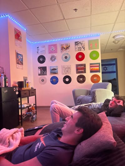 A rotated shot shows two individuals seated in a room with a casual, lived-in ambiance. Vinyl records adorn the wall, framed by a string of lights. Below, posters hang above a storage unit cluttered with various items. An Israeli flag is visible through the doorway. The atmosphere suggests a relaxed or domestic setting.