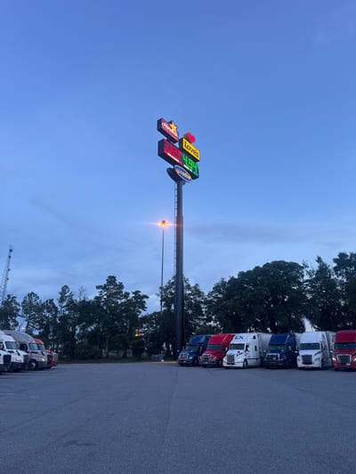 The image depicts a truck stop located in Lake City, United States, as indicated by the signage of "Hardee's" and "Loves." The scene is set at dusk, with a dim, twilight sky. A tall signpost prominently displays the logos of Hardee's and Loves, along with gas prices. The price for "Unleaded" is listed as $3.69, and "Diesel #2" is $4.94. Below these, "Speedco" is visible.

A row of semi-trucks is parked in the lot. These trucks vary in color, including dark blue, red, white with blue "EX" lettering, and white. The trucks are positioned in a line, facing towards the right side of the image. In the background, a cluster of trees forms a natural border, with some of their branches visible against the sky. A tall communication tower, with a lattice structure, is situated to the left of the scene. A street lamp with a bright light is positioned near the signpost, illuminating the immediate area. The ground of the truck stop is paved with asphalt. No people are visible in the image.