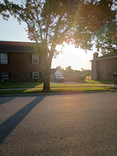 The image depicts a residential street scene in the city of Richmond, United States of America, during what appears to be late afternoon or early evening, as indicated by the long shadows cast by a large tree in the foreground. The sun is low in the sky, creating lens flare and illuminating the leaves of the tree.

On the left side of the image is a two-story brick building with multiple windows. The building has a dark roof and dark shutters on some of the windows. A grassy lawn with some decorative elements, including what appears to be a sunflower, is situated in front of this building.

A paved road occupies the bottom half of the image. The shadow of the prominent tree stretches across the road from left to right.

In the background, behind a wooden fence, a dark-colored SUV is parked. Further in the distance, more residential buildings and trees are visible, along with a water tower. A brick chimney from another building is also prominent in the background. The overall environment suggests a quiet suburban neighborhood. There are no people visible in the image.