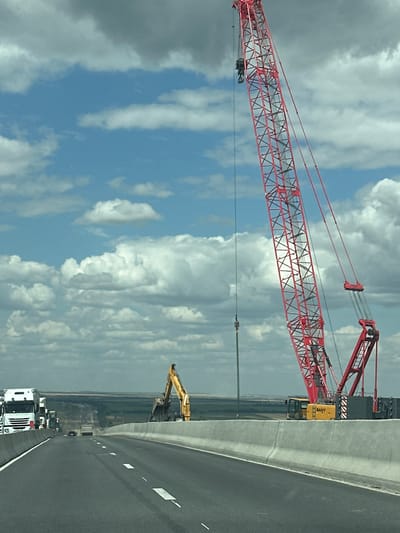 This image shows a construction site on a highway, likely a bridge under construction. A large red crane dominates the right side of the frame, with its boom extended upwards and to the right. A yellow excavator is visible in the middle ground, positioned behind a concrete barrier. Several white trucks are parked on the left side of the highway, suggesting ongoing work. The scene is set against a backdrop of a cloudy sky and a distant, flat landscape. The highway itself appears to be a multi-lane road with clear lane markings. The presence of construction equipment and barriers indicates that this is an active work zone. The specific location in Donleskhoz, Russia is not visually evident from the image itself, but the context suggests it is a remote or developing area. There are no people visible in the image.