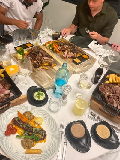 A high-angle shot depicts a dinner table laden with food and drinks, including plates of sliced meat, vegetables, sauces, and glasses. Two individuals are partially visible, one on the left wearing a dark green shirt, the other on the lower left wearing a white shirt with a patterned collar. Cutlery is arranged near each plate, and a blue glass bottle sits among the glasses in the center of the table. The overall setting suggests a meal in progress, but I have no basis to make a determination about its location in the city of Urbanització La Cometa, Spain.