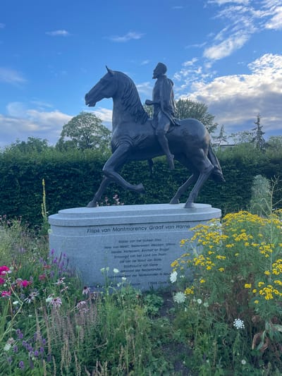 This is a vertical, eye-level, outdoor medium shot of a bronze equestrian statue. The statue depicts a man in historical attire riding a horse. The man wears a cloak and a hat, and his gaze is directed forward. The horse is captured in mid-stride, with its head slightly lowered. The statue stands on a light gray, cylindrical pedestal inscribed with text.
The pedestal's inscription, partially visible in Dutch, appears to be a memorial to "Filips van Montmorency Graaf van Horne," listing his titles and significant dates, including "1524" and "1568". The statue is situated in a garden setting, with lush green foliage and a variety of flowers surrounding the base. Yellow wildflowers are prominent in the foreground, interspersed with white and purple blossoms.
The background features dense green trees and a bright blue sky with scattered, wispy white clouds, suggesting a daytime setting, likely late morning or early afternoon, with pleasant weather. The overall impression is one of historical commemoration within a peaceful, natural environment. The scene evokes a sense of historical significance and quiet contemplation.