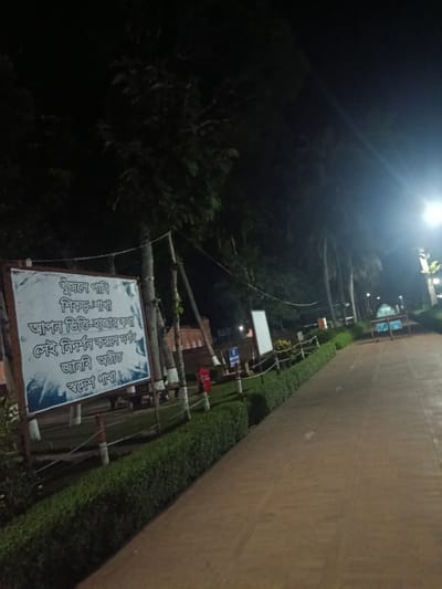The image shows a dimly lit pathway at night in Bagerhat, Bangladesh. A white sign with Bengali text is visible on the left, displaying a poem or message. The pathway is paved with reddish-brown material and lined with manicured green hedges on the left. Trees with dark foliage are present on both sides of the pathway, with some branches extending over the scene. A street light illuminates a portion of the sky and pathway on the right side of the image. There are no people visible in the frame. The overall atmosphere is quiet and serene.
