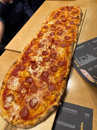 A long, rectangular pizza, approximately one meter in length, is presented on a light wooden paddle board on a light-colored wooden table. The pizza is topped uniformly with tomato sauce, melted mozzarella cheese, and circular slices of pepperoni (salame piccante). The crust is golden-brown. The pizza has been pre-cut into numerous rectangular portions, arranged in two parallel rows. To the right of the pizza, two black menus are visible; one is titled "Bevande" (Beverages) and lists items with prices in Euros, while the other shows "Birra alla Spina" (Draft Beer) with volumetric options and corresponding Euro prices. In the blurred upper left background, parts of individuals are visible, suggesting other patrons in a dining environment. The scene is situated within a dining establishment in Foligno, Italy.
