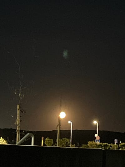The image depicts a night scene in an urban environment. Several streetlights illuminate the area. A row of trees or bushes lines the right side of the image. In the dark sky, there is a faint greenish anomaly. Based on the provided context, the location is likely Mensdorf, Luxembourg.