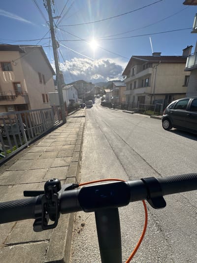 A sunny day in Borino, Bulgaria, captured from the perspective of someone riding an electric scooter down a quiet street. The scooter's handlebars and control panel are in the foreground. The street extends into the distance, flanked by buildings of varying heights and architectural styles, typical of a small European town. A parked car is visible on the right side of the street, partially cut off by the image frame. Power lines crisscross the sky, contrasting with the blue sky and scattered clouds. A contrail from a high-flying aircraft is visible in the upper left corner. The sun shines brightly, creating strong shadows on the street and buildings. The overall impression is one of a peaceful residential area on a clear, bright day.