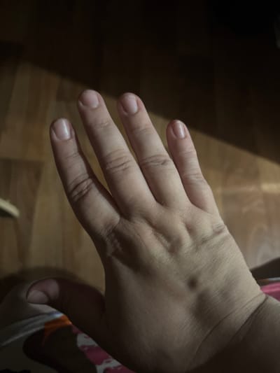 A close-up shot focuses on the back of a person's left hand, with the fingers extended and slightly splayed. The hand is positioned against a dark background, with the lower half of the image revealing a wooden floor bathed in warm, artificial light. The light creates distinct shadows and highlights on the floor, suggesting a domestic indoor setting.
The skin on the back of the hand appears to be fair-toned, with visible lines and creases indicating age or natural skin texture. Four fingers are clearly visible, each with a naturally trimmed fingernail. The thumb is mostly out of frame, with only a hint of its base visible near the right edge of the image. A small, raised bump is noticeable on the back of the hand, between the knuckles of the index and middle fingers.
To the right of the hand, a colorful patterned fabric is partially visible, suggesting the person might be wearing clothing with a vibrant design, possibly pajamas or casual wear. The overall lighting is dim, with the primary light source coming from the side, illuminating the hand and a portion of the floor. The dark background and the warm glow on the floor evoke a sense of quietude, perhaps late evening or early morning. There is no visible text in the image. The scene does not depict any specific activity other than the resting of the hand.