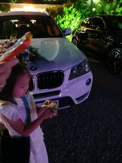 A young girl, with dark hair and a white dress, stands in front of a white BMW SUV, holding a small, golden decorative object. She is also holding a red and orange object that resembles a small toy or decoration. The car is adorned with a bouquet of purple and white flowers on its hood. To the right of the BMW, a black car, possibly an Alfa Romeo, is parked. The scene is set at night, with artificial lighting illuminating the cars and the surrounding green foliage. The ground appears to be a gravel surface. The presence of the BMW suggests a formal occasion, possibly a wedding or a special event, taking place in Kırıkkale, Turkey.
