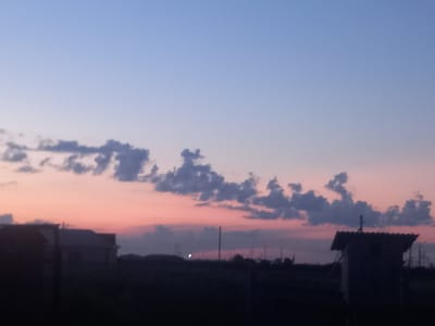 A twilight scene in Қозыкеткен ауылдық округі, Kazakhstan features a soft gradient sky transitioning from pale blue at the top to a peachy pink hue near the horizon. Scattered, dark grey clouds are visible in the mid-sky. The foreground is dominated by a dark silhouette of the landscape, with indistinct structures suggesting rural buildings and possibly utility poles. On the right side of the image, a small, dark, shed-like structure with a sloped roof stands out against the darker background. The overall impression is a quiet, rural evening at dusk.