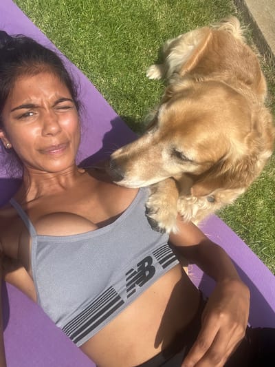 In Culver City, United States of America, a young woman is lying on a purple yoga mat outdoors on a sunny day. She has dark hair pulled back and is wearing a grey sports bra. A golden retriever is lying next to her, with its head resting on her chest. The background is a grassy lawn. The woman appears to be squinting into the sun.