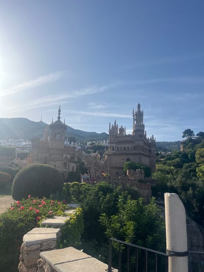 The image shows a panoramic view of a castle-like structure with intricate spires and stonework, set against a backdrop of lush green trees and mountains. The sky is a clear, bright blue with wispy white clouds, suggesting a sunny day. The time of day appears to be late afternoon, as indicated by the warm, soft light casting long shadows and creating a slight haze in the distance.

The castle, located in Benalmádena, Spain, is the prominent subject. It features multiple towers, turrets, and decorative architectural elements that give it a fairytale-like appearance. Greenery surrounds the structure, with trees and bushes forming a dense canopy on the hillsides and climbing up some of the castle's walls. In the foreground, there are hints of manicured gardens, with a rounded bush and blooming pink flowers adding color to the scene. Stone elements, possibly part of a pathway or wall, are also visible.

While the image is primarily focused on the architecture and natural landscape, small groups of people can be seen in the distance, suggesting that the location is a tourist attraction. They appear to be walking around and observing the castle. The overall mood is serene and picturesque, showcasing the beauty of the castle and its natural surroundings bathed in the golden light of the sun. There is no visible text in the image.