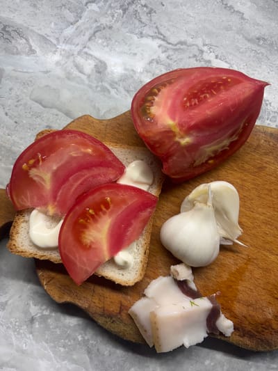 This media file shows an overhead view of a wooden cutting board placed on a marble-patterned surface. On the cutting board are several food items. Two large slices of ripe red tomato are visible, one in the upper left quadrant and the other on a slice of toasted bread in the lower right. The tomato slices are juicy with visible seeds. The bread is toasted and appears to have mayonnaise spread on it, with two tomato slices arranged on top. To the right of the large tomato slice, there are two cloves of garlic. Next to the garlic, a piece of cured pork fatback, known as salo in Ukraine, is present. The salo is white with a thin layer of dark meat and some green herbs, likely dill, sprinkled on top. The overall scene suggests the preparation of a simple meal or snack. The lighting appears to be natural, casting soft shadows, and there is no indication of the time of day or weather. The setting is likely a kitchen counter or a similar preparation area in Pereiaslav, Ukraine, given the context. No people are visible in the frame, and there is no visible text.