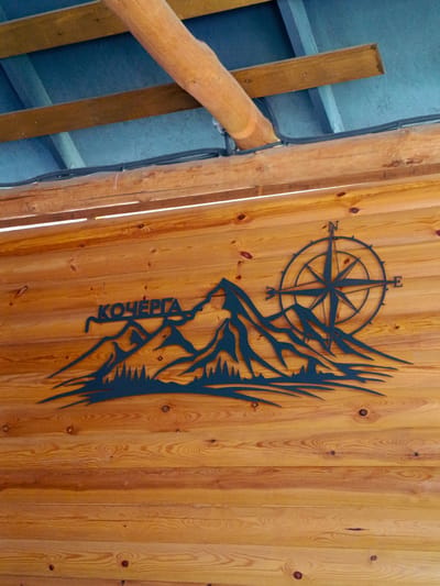 A dark metal silhouette depicting mountains and a compass rose is mounted on a wooden wall. The silhouette is on the left side of the image, with the mountains forming a jagged line across the lower portion and a compass rose on the right. The mountains are dark blue, with sharp peaks and a forest line at their base. The compass rose is also dark blue, with the letters 'N' and 'E' visible. The wooden wall is made of horizontal planks with visible grain and knots. Above the wall, there are wooden beams and blue material, possibly a roof. This scene appears to be outdoors, possibly on a building or structure. The location is identified as Kok Jar, Kyrgyzstan.