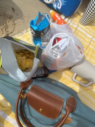 The scene depicts a collection of items resting on a yellow and white striped towel spread across a sandy beach surface in South Padre Island, United States of America. No individuals are visible.

In the foreground, a light blue/teal nylon tote bag with brown leather handles and a brown leather flap featuring a gold-colored snap is prominently displayed. Above the tote bag, two open bags of potato chips are visible: one large bag with a green-and-white label containing numerous potato chips, and a smaller blue-and-black bag with a "Nutrition Facts" label and some chips remaining. Adjacent to these chip bags, a transparent plastic grocery bag contains additional packaged food items, with a white label displaying "Nutrition Facts" and the number "140".

Other objects include an orange tube of sunscreen labeled "ULTRA" partially visible to the upper right, a blue and white patterned fabric bag (likely a beach bag) also partially visible in the background, and a white insulated mug with a handle positioned on the lower right of the towel. A dark brown plastic bottle is partially visible near the blue chip bag. The environment is well-lit by sunlight, casting some shadows.