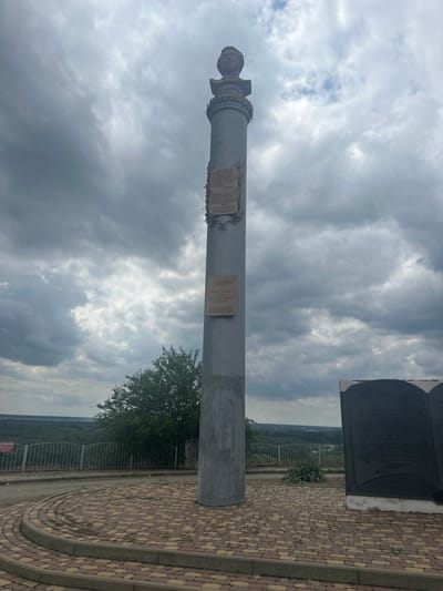 The media file depicts a monument in Novokubansk, Russia, set against a dramatic sky filled with dark, heavy clouds. The main subject is a tall, grey, cylindrical monument, possibly a column, topped with a bust of a person. There are at least two plaques attached to the monument, featuring text that is not entirely legible due to the image quality and angle. The monument stands on a paved area with a distinctive yellow and reddish brick pattern laid in concentric circles.

To the right of the monument, a large, dark, book-shaped structure is visible, also with inscriptions. A metal fence runs along the edge of the paved area, overlooking a landscape of rolling hills covered in dense green trees. The time of day appears to be overcast, perhaps late afternoon or early evening, given the dim lighting and the ominous clouds. The overall mood is somber and dramatic, enhanced by the weather and the imposing presence of the monument. No specific activity or event is taking place; the scene appears to be a still observation of the monument and its surroundings. There are no other people visible in the frame.