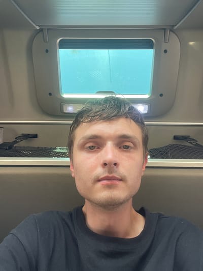 The image shows a close-up, selfie-style shot of a man in what appears to be a vehicle or train cabin. The man has short, dark hair, light skin, and light-colored eyes. He is wearing a black t-shirt. In the background, there is a window with a blue tint, and overhead compartments with what appear to be storage nets. There is also an overhead light fixture. The overall environment suggests an interior space within a mode of transportation. The location is identified as Brachterbeek, Netherlands.
