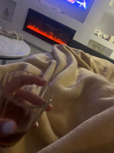 A person's hand holds a clear glass in front of a soft, beige blanket. An electric fireplace with a vertical orange glow is visible on the left side of the frame. A white, round table and a partially visible towel are near the fireplace. In the background, a television screen and decorative shelves are visible, completing the cozy indoor scene.