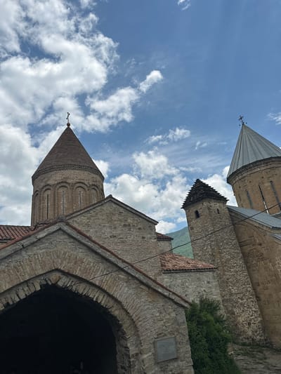 The image depicts the Ananuri fortress complex located in Shalikiantkari, Georgia. The complex consists of several stone structures, including churches with conical and conical-domed roofs topped with crosses. The architecture is characterized by intricate stonework and visible brick detailing. The buildings are set against a backdrop of a bright blue sky with fluffy white clouds and hints of green mountains in the distance. The foreground features an arched entryway made of stone and brick. There are no people visible in the image.