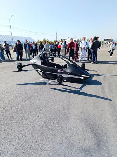 In Korsakov, Russia, on a paved surface, a black, two-seater, drone-like vehicle with four rotors is parked. The vehicle has the text "X-SKY" visible on its side. A group of approximately 20-30 individuals, appearing to be spectators and participants, are gathered around the vehicle, some observing it and others interacting amongst themselves. The environment suggests an outdoor event or exhibition, with a clear blue sky overhead and a large white structure in the background. The people are casually dressed, with some wearing athletic attire. The lighting indicates it is daytime, with shadows cast by the vehicle and people.