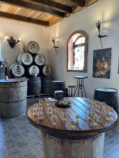 The image depicts the interior of a rustic establishment, possibly a distillery or tasting room, located in the Літвенскі сельскі Савет, Belarus. The space is characterized by aged wooden barrels stacked in various locations, serving as both display and functional elements. Dominating the foreground is a large, round wooden table, possibly repurposed from a cable spool, set with numerous small glasses of amber liquid, suggesting a tasting session. A plate of small green appetizers is also visible on this table.
The walls are whitewashed, contrasting with the dark wooden beams and planks of the ceiling, which also features exposed wooden joists. A large arched window with brick detailing is visible on the right side of the room, letting in natural light and revealing greenery outside. Above the window, a painting hangs on the wall depicting a historical scene with a man in traditional attire. Wall-mounted torch-like sconces with flickering light fixtures are present on the walls, adding to the antique ambiance.
Several tall, narrow tables with matching stools are placed near the window, offering additional seating. In the background, more wooden barrels are visible, some quite large, contributing to the overall theme of barrel-aged products. The floor is tiled with a patterned design. No people are present in the scene, giving the impression of a quiet moment or a setup prior to an event. The lighting is a mix of natural light from the window and the artificial light from the wall sconces.