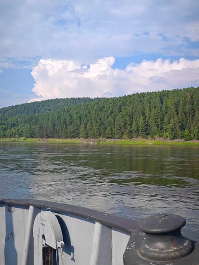 A river flows in the foreground with a dense, green pine forest covering a sloping hillside in the background. The sky above is a mix of blue and white clouds, with a large, fluffy cloud formation on the left. The viewpoint appears to be from a boat, as the edge of a grey deck railing and a mooring cleat are visible in the lower part of the image. The location is the river in Beregovaya-Podemnaya, Russia.
