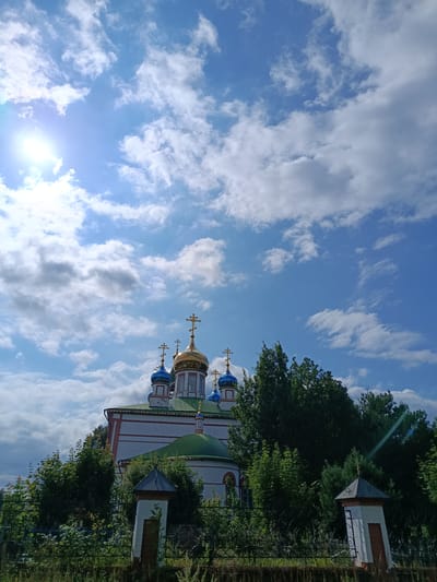 The image shows the Church of the Intercession in Staryi Pokrov, Russia, under a partly cloudy blue sky. The church has a white facade with red trim, a green roof, and multiple golden domes topped with crosses, as well as blue domes. Tall green trees surround the church. In the foreground, a black metal fence runs across the bottom of the image, with two small architectural elements on posts to the right of the frame. The sun is visible in the upper left quadrant, casting a lens flare. The scene depicts a place of worship and its surrounding natural environment.