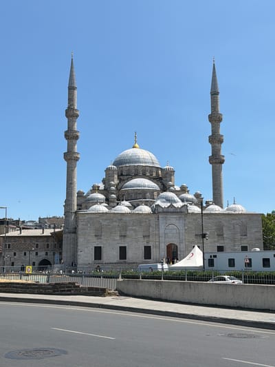 The image shows the Yeni Cami (New Mosque) in the Eminönü district of Fatih, Istanbul, Turkey. The grand mosque features a central dome, flanked by smaller domes and supported by stone minarets. The building is constructed from light-colored stone, and its architectural details are intricate. In front of the mosque, there is a street with a sidewalk and a low concrete wall. Beyond the wall, a metal fence encloses an area with some people, a white tent with a red crescent logo, and a white mobile unit. Trees are visible on the right side of the image, and the sky is clear and blue.