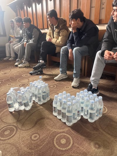 A group of at least six young males are seated on chairs, facing to the left, with four stacks of bottled water positioned on the patterned carpet in front of them. The location is likely inside a building in Charentsavan, Armenia. The men are dressed in casual clothing including jeans, sweatpants, jackets, and sneakers. Some appear to be looking down. The water bottles are packaged in plastic wrap. The carpet has a brown background with a circular pattern. The walls appear to be made of wood panels.
