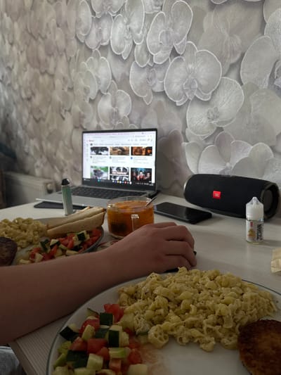 The image shows a table setting with various items. A person's arm is visible near a plate of food consisting of pasta, a patty, and a tomato-cucumber salad. Next to the plate, there is a sandwich and a glass of orange liquid. A laptop displays a webpage with thumbnails of videos. To the left, a black JBL speaker and a bottle labeled "NARCOZ" are placed. The table surface is white. The wallpaper features a white floral pattern. The location in the city of Shirkovo, Russia, cannot be confirmed from the provided image.