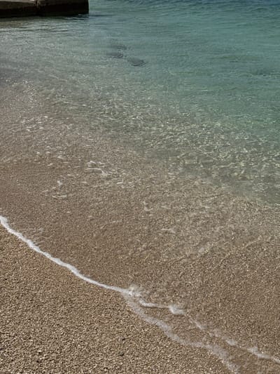 The image shows a close-up view of a sandy beach in Dugi Rat, Croatia. The shoreline is marked by a thin line of white foam, the receding edge of a wave, and the water is clear, showing the pebbly bottom. The water is calm, and there are no people or objects visible in the image. The beach is made up of fine-grained sand and small pebbles. The water is a clear, pale turquoise, with ripples caused by the gentle current. The sand is a light brown color, and the pebbles are a mixture of brown and gray. The image captures a peaceful and tranquil scene.
