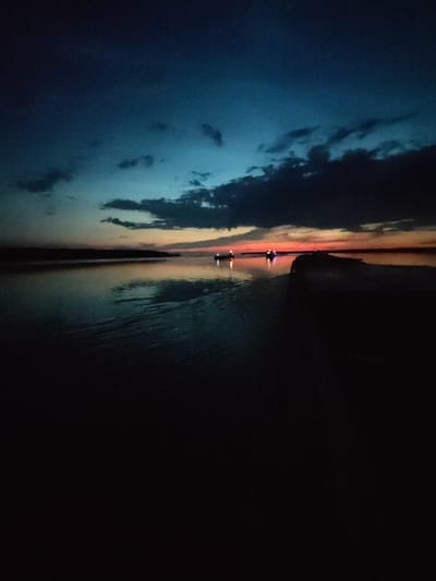 The image captures a serene twilight scene on the water, possibly the Gulf of Finland, near Kamenniki, Russia. The sky transitions from a deep, dusky blue overhead to hues of orange and pink near the horizon, indicating the last moments of sunset. Dark, scattered clouds drift across the upper sky.
The water in the foreground is dark and reflective, showing subtle ripples. In the mid-ground, two small boats with lights illuminated are visible on the water, suggesting they are either fishing or transiting the area. A long, dark, man-made structure, possibly a pier or breakwater, extends into the water from the right side of the frame, creating a stark contrast with the lighter horizon. The far shore is a dark, indistinct silhouette against the colored sky. The overall impression is one of quietness and solitude as night descends.
