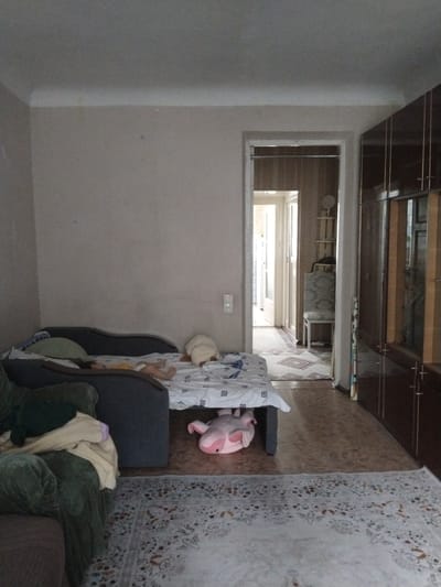 The image shows an indoor scene, likely a living room or bedroom, in a dwelling located in Ötegen batyr, Kazakhstan. In the foreground, a large, dark gray sofa is partially visible on the left, with a green armchair in front of it draped with blankets. A bed with white bedding featuring a black geometric pattern is positioned in the center of the room. On the bed, a person is lying down, wearing a yellow top and what appears to be a diaper. A large, light-colored stuffed animal is on the right side of the bed. Another pink stuffed animal is lying on the floor underneath the bed. To the right of the bed, a doorway opens into another room, where a chair with a patterned upholstery is visible. Further to the right is a tall, dark wood cabinet with multiple doors and drawers. The flooring consists of a light brown patterned linoleum and a cream-colored rug with a floral pattern in the foreground. The walls are painted a neutral color, and the ceiling is visible at the top of the image. Natural light enters the room from the doorway in the background, illuminating the scene.