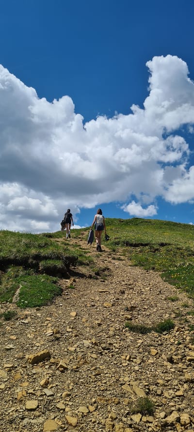 The image shows two people hiking on a rocky mountain path with green grass and yellow wildflowers on a sunny day. The sky is blue with large white clouds. One hiker is in front, wearing a dark shirt and shorts, and the other is behind, wearing a white tank top and shorts. They appear to be ascending the mountain. The scene suggests a recreational activity in a natural mountain environment, likely during daylight hours given the bright sunshine and blue sky. The terrain is uneven and appears to be a trail. There is no visible text in the image. The overall impression is one of a pleasant outdoor excursion.