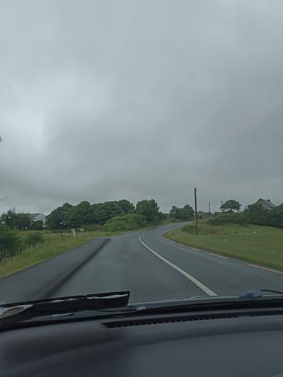The image, captured from the interior of a vehicle, shows a grey, overcast sky. A paved road with white and yellow lines curves to the left. On the left side of the road, there is a white house with a dark roof, surrounded by greenery. Further down the road, on the right, are more trees and a house with a dark roof and a chimney. There are some sheep visible in the grassy field on the right. The foreground shows the dashboard and windshield wipers of the vehicle. This scene is consistent with the landscape of Ranafast, Ireland, which is known for its rural, coastal setting with scattered houses and rolling hills under often cloudy skies.
