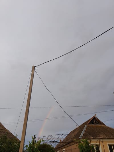 A wide shot captures an overcast sky over the village of Parkany, Moldova. A tall, grey concrete utility pole stands prominently on the left, supporting multiple black wires that stretch across the frame. In the distance, a faint rainbow arcs through the sky. The lower portion of the image reveals the rooftops of houses. The roof on the left is made of dark, corrugated material, while the larger roof on the right is covered in a similar weathered material and features a triangular attic window. Overgrown green vines are visible in the foreground, climbing on a metal structure. The scene is devoid of any people or animals.