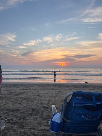 A beach scene during sunset with orange hues. The sun is a small, bright orb on the horizon. Silhouetted figures stand in the water. Gentle waves roll onto the sandy shore. A blue beach chair occupies the right side, with a blue and white towel draped over its arm. The sand is coarse and dark. The sky features wispy clouds, transitioning from blue to orange near the sunset.