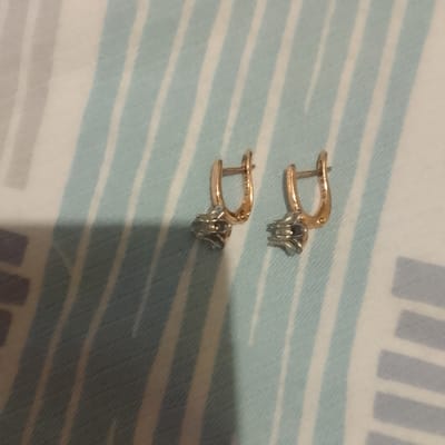 Two gold earrings with small diamond-like stones are placed on a striped fabric surface. The fabric has alternating light blue and white stripes. The earrings are in focus and are the main subjects of the image. No specific location in Almaluu, Kyrgyzstan is discernible from the image.