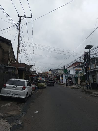 A street scene under a cloudy sky features a white car parked on the left, partially obstructing the sidewalk. A light green minivan and a black SUV are further down the road. Overhead, a dense web of power lines crisscrosses the sky, supported by utility poles. Buildings line both sides of the street, with various shops and businesses visible. A person is working on a utility pole on the right. The road surface is asphalt, showing signs of wear and patches.