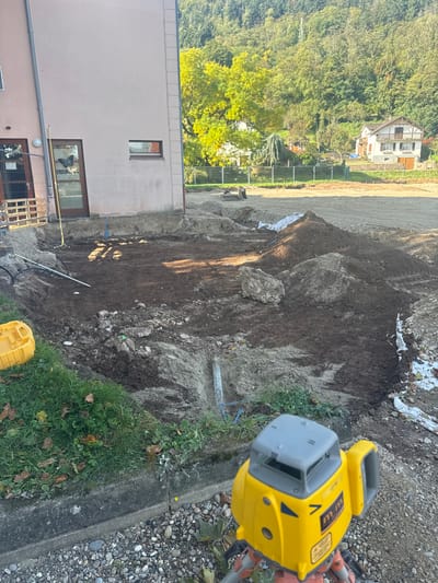 The image displays an active construction site in Châtenois, France, under clear daylight conditions. In the immediate foreground, a yellow and grey laser level, mounted on a tripod, is positioned on a gravel surface adjacent to a concrete curb and a strip of green grass. A large, irregular excavation pit dominates the midground, exposing layers of dark topsoil and lighter subsoil. Several white PVC pipes and black utility lines are visible within and around the excavated area. Piles of disturbed earth and rocks are situated next to the pit. A yellow plastic container rests on the grass verge to the left of the excavation. Abutting the left side of the pit is a light-colored building, possibly pale pink or beige, featuring a dark brown door adorned with a large butterfly decal and a single rectangular window. A yellow measuring tape or line extends from near the building's door into the excavation. In the background, beyond the immediate construction zone, a wide expanse of leveled, disturbed earth is visible, leading to a distant fence line. Behind this, a light-colored, multi-story house with dark trim and a prominent balcony is situated, backed by a densely wooded hillside exhibiting varied green and yellow autumn foliage. No individuals or active construction machinery are visible in the scene.