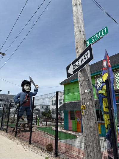 The image displays an outdoor scene under a clear, bright blue sky, suggesting daytime. A sturdy wooden utility pole stands prominently on the left, featuring two main signs. A green street sign attached to the pole clearly reads "N STREET." Below it, a black and white "ONE WAY" sign with a directional arrow points to the right. Yellow numbers "69" are also visible on the pole. Several dark power lines stretch across the upper portion of the sky.
To the right of the utility pole, a tall black mesh fence separates the foreground from a colorful area behind it. A large, anthropomorphic pirate statue, wearing a black hat, a white face covering or mask, a blue jacket, and a red sash, stands holding a raised cutlass behind this fence. The area beyond the fence appears to be a miniature golf course, with visible green turf and red-brown pathways.
In the background, several buildings are painted in vivid, contrasting colors such as lime green, orange, pink, and yellow, characteristic of a recreational or tourist venue. One building has a small sign featuring an ice cream cone graphic. Above the buildings and fence, colorful fabric banners or kites are partially visible, adding to the lively atmosphere. The immediate foreground includes a concrete sidewalk and a strip of gravel beside the fence. The scene conveys the ambiance of a bustling tourist or entertainment area.