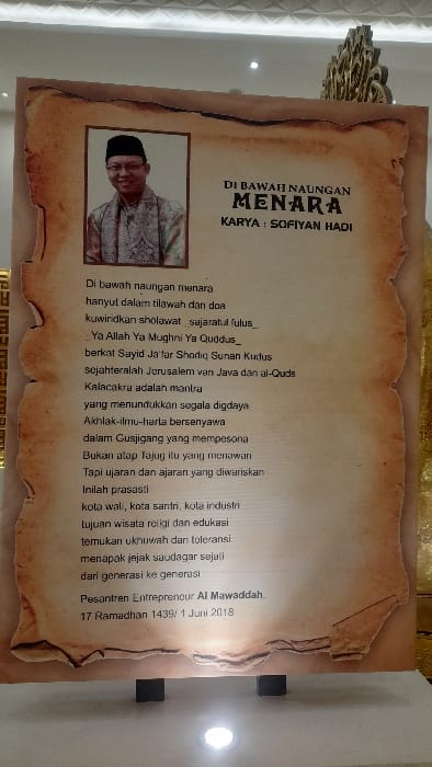 This image, captured indoors in Glantengan, Indonesia, displays a large, vertical standee designed to resemble an antique, frayed parchment scroll. The standee, predominantly sepia-toned with jagged, worn edges, measures approximately human height and is supported by two small, black rectangular bases on a light-colored floor.
A central photograph on the upper left of the standee shows a smiling man in a black songkok (cap) and a patterned light-colored shirt or robe. To the right of the photograph, black Indonesian text is visible, with the prominent title "DI BAWAH NAUNGAN MENARA" (Under the Shadow of the Tower) and "KARYA : SOFIYAN HADI" (Work by: Sofiyan Hadi). Below this, a lengthy text, likely a poem or prose, begins with "Di bawah naungan menara..." and concludes with "Pesantren Entrepreneur Al Mawaddah. 17 Ramadhan 1439/ 1 Juni 2018."
The surrounding environment consists of a light-colored floor reflecting light from a bright, circular recessed lamp positioned directly in front of the standee at floor level. The background features light-toned walls or panels, with a textured ceiling visible at the top, equipped with multiple recessed light fixtures. A decorative, ornate golden structure is partially visible on the far right, behind the display board. No individuals are present or engaged in direct interaction within the frame.