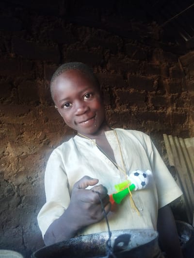 A young child smiles at the camera while standing in what appears to be an indoor space with a rough, textured wall. The child wears a light-colored shirt and holds a noisemaker with a soccer ball attached. A string hangs around the child's neck, likely connected to the noisemaker. Cooking utensils are visible in the foreground. The environment suggests a simple or rural setting.