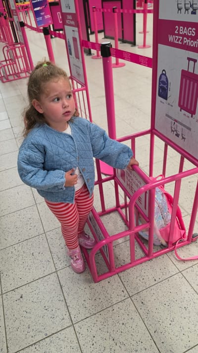 The image depicts a young girl standing next to pink metal frames, likely luggage sizers or queue barriers, in what appears to be an airport setting. The girl has light brown, curly hair and is wearing a light blue quilted jacket, red and white striped leggings, and pink shoes adorned with a butterfly design. She is looking upwards with a curious or thoughtful expression, holding onto one of the pink frames.
A small pink backpack with iridescent sequins is placed inside one of the luggage sizers, suggesting a bag is being checked for size compliance. The floor is made of light-colored, speckled tiles.
Visible text on the pink signs and branding includes "2 BAGS WIZZ Priority" and dimensions like "40 cm", "30 cm", and "20 cm", indicating carry-on luggage requirements for Wizz Air. The WIZZ logo is also visible on pink straps and other signage. Partially visible text on another sign reads "ONE YOUR TROLLEY BAG", likely instructing passengers on bag placement.
The bright pink color scheme of the airport equipment is a prominent feature, strongly associating the scene with Wizz Air, often seen in airports like Luton, United Kingdom. The lighting suggests an indoor setting.