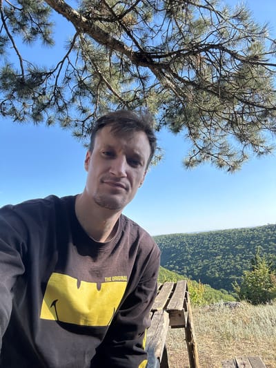 A man with short brown hair and stubble is taking a selfie outdoors. He is wearing a dark brown long-sleeved shirt with a large yellow smiley face graphic and the words "THE ORIGINAL" above it. He is seated on a wooden bench, with a lush green, hilly landscape stretching out behind him under a clear blue sky. Pine branches hang overhead, partially obscuring the sky. The scene appears to be a scenic overlook, likely in the vicinity of Nihyn, Ukraine, given the geographical context.