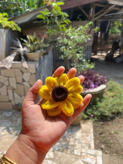 An extreme close-up shot shows a person's hand holding a yellow and brown pipe cleaner flower. The flower is in focus, while the background is blurred, showing a residential outdoor environment with some greenery and structures. The scene is likely in Beuringen, Indonesia, given the surrounding vegetation and architecture.