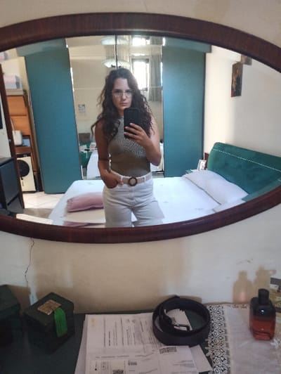 An individual is depicted in a full-length, oval-framed mirror selfie taken indoors, specifically a bedroom in San Marzano sul Sarno, Italy.
The subject is a female with long, dark, wavy hair, wearing eyeglasses. She is dressed in a light-colored, ribbed, sleeveless top and light-colored high-waisted shorts or trousers, secured with a light-colored belt featuring a circular buckle. Her left hand holds a black smartphone to capture the image, while her right hand is placed into a pocket.
The reflection in the mirror reveals a room with light-colored walls, one displaying a visible vertical crack below the mirror's frame. A bed with a dark green upholstered headboard, white sheets, a white pillow, and a pink cylindrical pillow is visible. To the left of the bed, two teal-colored sliding doors are present, likely part of a wardrobe. A window is reflected further left, allowing natural light into the room. A multi-tiered ceiling light fixture is also reflected overhead. Above the bed's headboard, a small, dark-toned picture or icon is affixed to the wall.
The foreground, directly beneath the mirror, is a dark-surfaced table or dresser. On its left, a dark green rectangular box with gold accents and a green tassel is situated. In the center, several white documents or papers, including one with a visible QR code, are stacked. To the right, a dark brown leather belt is coiled, resting on a white mat with a patterned design. Adjacent to the belt, a reddish-orange glass bottle, possibly containing perfume, is present.