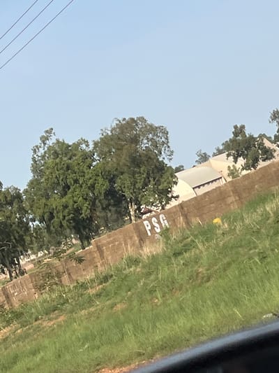 The image shows a dry-stone wall with the word "PSG" spray-painted on it in white. Behind the wall, there are several trees and a building with a light-colored roof. The ground in front of the wall is covered in green grass. The image appears to have been taken outdoors during the daytime, with a clear blue sky. The location is identified as Rahwol Chom, Nigeria.