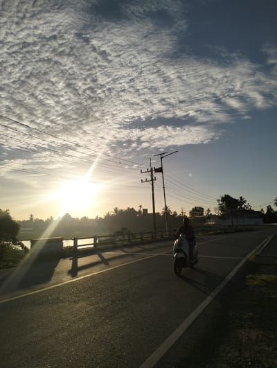 The image shows a sunlit street scene in Teungkluet, Indonesia. A person is riding a white scooter on a paved road with a solid white line marking the edge. The sun is low in the sky, casting long shadows from a bridge railing and utility poles. Overhead power lines crisscross the sky, which is filled with patterned clouds. Trees and vegetation line the side of the road near the bridge. A building is visible in the distance.
