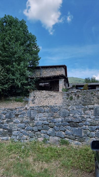 The image shows a rustic stone building with a sign that reads "HOSTAL DE LA ROVIRA RESTAURANT". The building appears to be a restaurant or an inn, likely located in a rural area given the surrounding stone walls, trees, and vegetation. A portion of a car mirror is visible in the lower right corner. The sky above is blue with scattered white clouds, and a green hillside is visible in the background. The overall scene suggests a peaceful and natural setting. The building is constructed from rough-hewn stones, with a tiled roof. A covered porch or veranda is visible in front of the main structure.