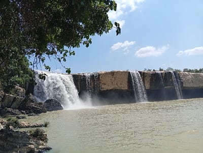The image depicts a wide waterfall located in Đức Lập, Vietnam, under a clear blue sky with sparse white clouds. Water cascades over a broad, stratified rock formation, creating a significant main fall on the left and several smaller, distinct streams to the right. A large pool of murky, greenish-brown water collects at the base of the falls, extending into the foreground. The left bank features rugged, dark rock formations, appearing columnar in places, with dense green vegetation and tree branches overhanging from the upper left. The top edge of the waterfall cliff displays exposed tan and dark rock layers. No individuals, human-made objects, or specific actions are discernible within the frame.